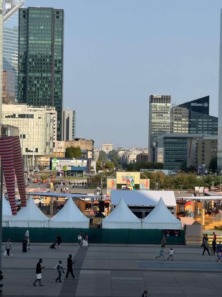 Elevated view of the La Défense esplanade looking towards the Arc de Triomphe, featuring a temporary event setup with white tents and a colorful "Garden Parvis" sign, surrounded by modern skyscrapers and pedestrians on the aging concrete deck.