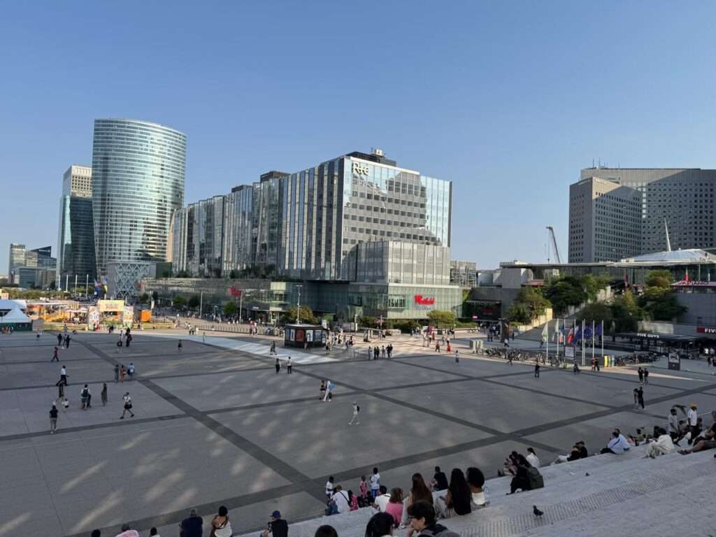 Wide elevated view from the Grande Arche steps overlooking the La Défense concrete esplanade. A diverse crowd sits in the foreground, while the Westfield shopping center, featuring a prominent American fast-food storefront, dominates the right side of the aging plaza under a clear blue sky.