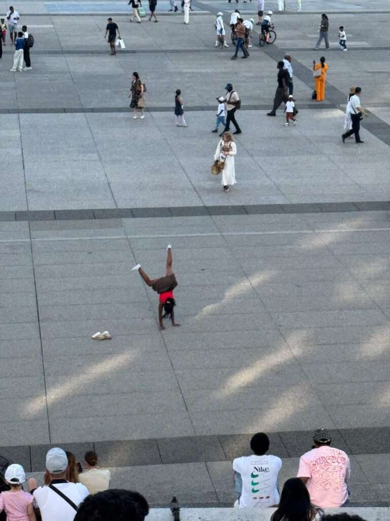 Elevated view of a young girl doing a handstand on the paved concrete esplanade of La Défense, while casually dressed people sit on the steps in the foreground and diverse pedestrians stroll in the background.