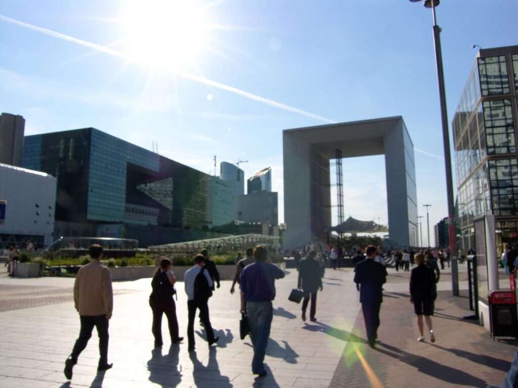 Silhouette of the Grande Arche and modern glass buildings against a bright, sunlit sky, with business professionals walking across the tiled plaza in the foreground.