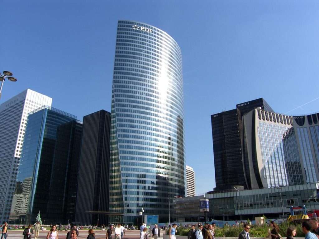The cylindrical glass EDF tower dominating the skyline, with businessmen in suits walking across the sunlit concrete and brick-paved plaza in the foreground.