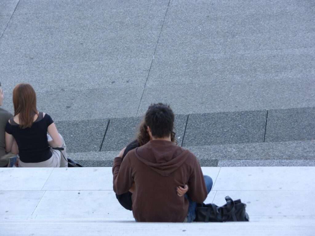 View from behind of a young couple sitting closely together on the white marble steps of the Grande Arche, with a black bag resting beside them on the stark concrete.