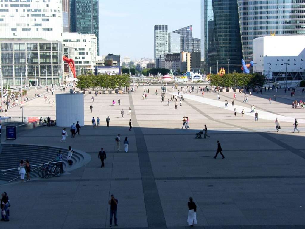 Wide, elevated view of the La Défense esplanade covered in large square concrete tiles, with scattered pedestrians walking towards the distant historical axis.