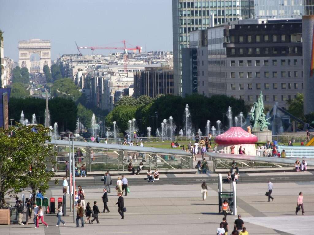 The La Défense esplanade looking towards the Arc de Triomphe, featuring active water fountains, small event structures, and scattered visitors on the concrete deck.