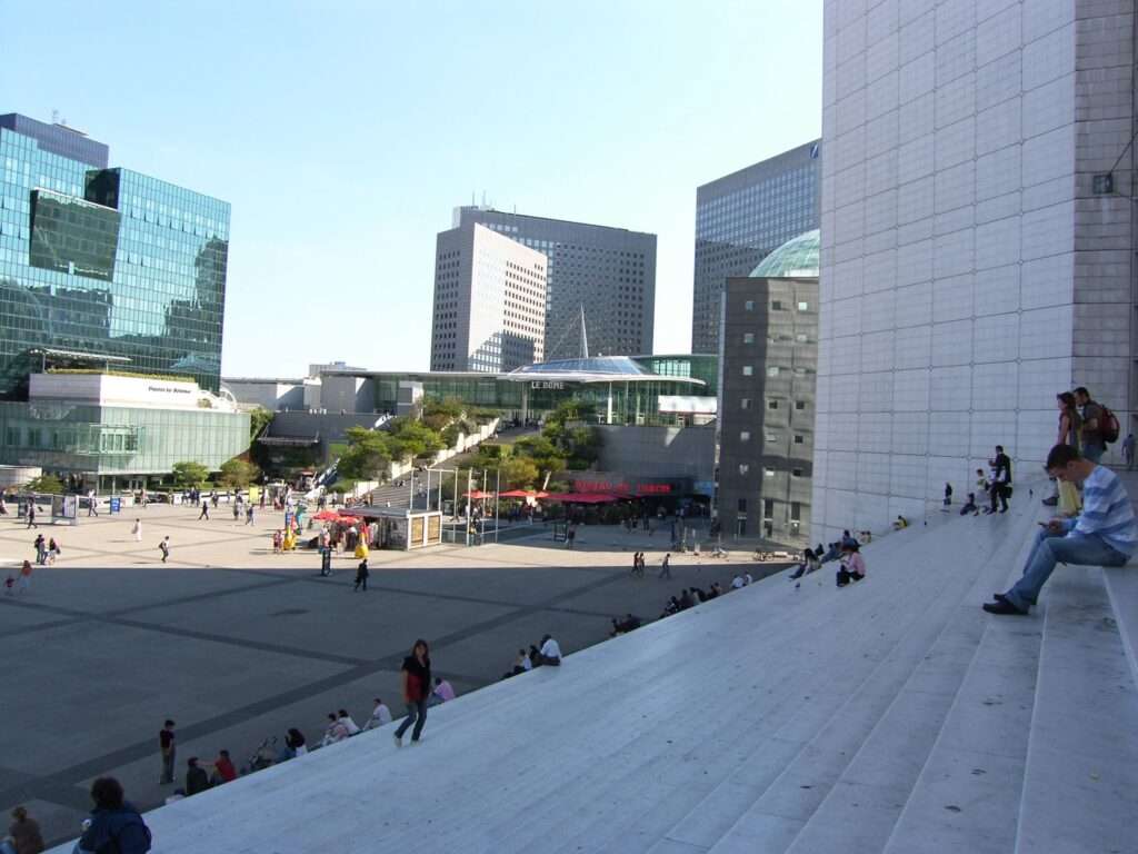 View looking down from the white steps of the Grande Arche onto the main plaza, with a few people resting on the stairs and the Le Dôme shopping center in the distance.
