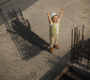 ArchUp satirical photograph showing a smiling toddler dressed in a hard hat and safety vest standing on a raw concrete construction site with arms raised next to steel rebar casting a long shadow