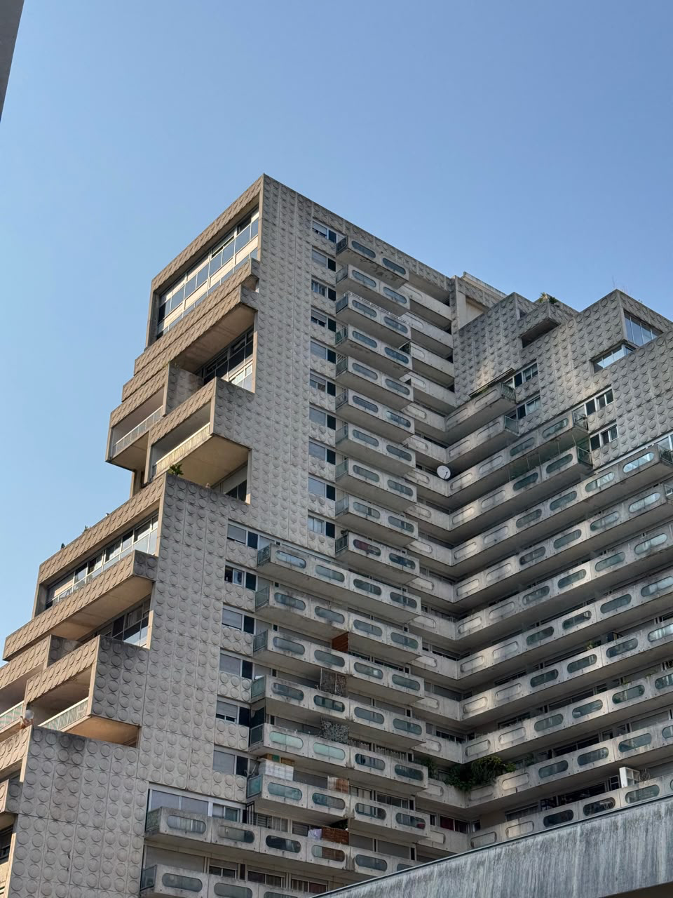 Looking up at a massive Brutalist residential building made of raw concrete, featuring a stepped design, pill-shaped balcony openings, and distinctive circular patterns embossed on the exterior panels.