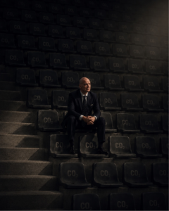 ArchUp AI-generated image depicting FIFA President Gianni Infantino sitting alone on dark concrete stadium bleachers where every empty seat is prominently stamped with the text CO2 under dramatic cinematic lighting