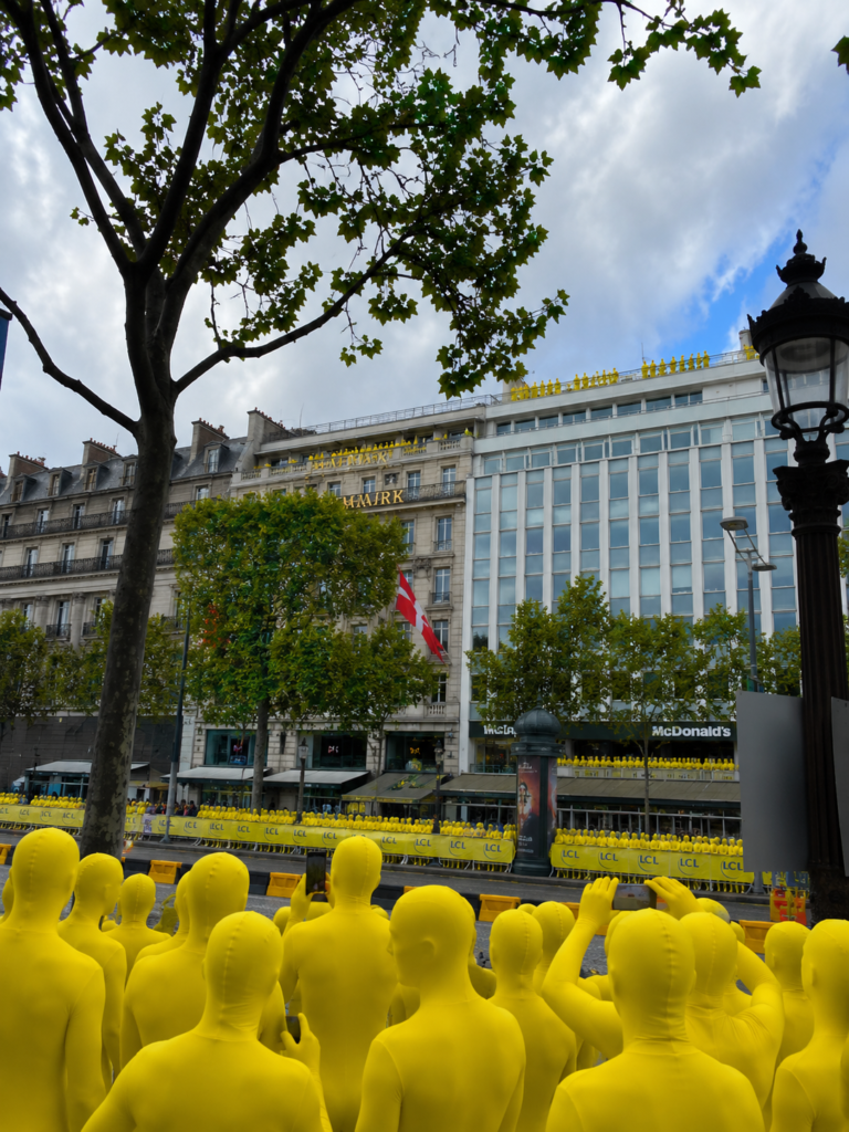 AI-generated conceptual image of a Parisian street scene where a massive crowd of featureless, bright yellow humanoid figures acts as spectators. They are gathered densely in the foreground holding up phones, lined behind yellow barricades along the boulevard, and standing across the rooftops of classic Haussmann and modern commercial buildings.