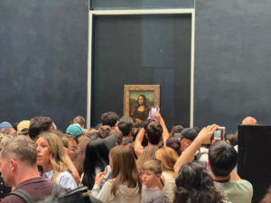 A dense crowd of tourists holding up smartphones to photograph the Mona Lisa painting, which hangs centered on a large dark wall inside the Louvre Museum.