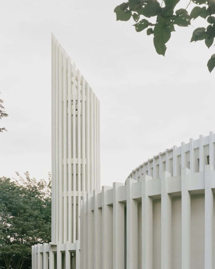 Curved semi-outdoor corridor at Santuario de La Salle flanked by a smooth white wall, vertical concrete pillars, and a minimalist hanging light fixture.