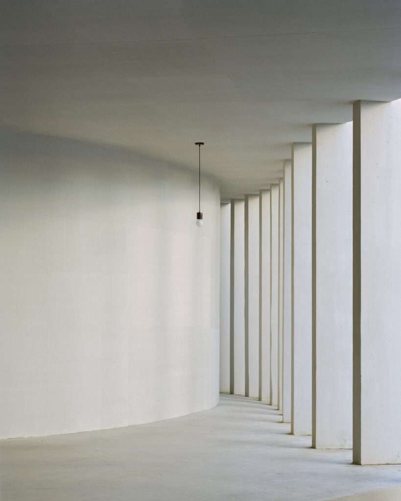 Curved semi-outdoor corridor at Santuario de La Salle flanked by a smooth white wall, vertical concrete pillars, and a minimalist hanging light fixture.