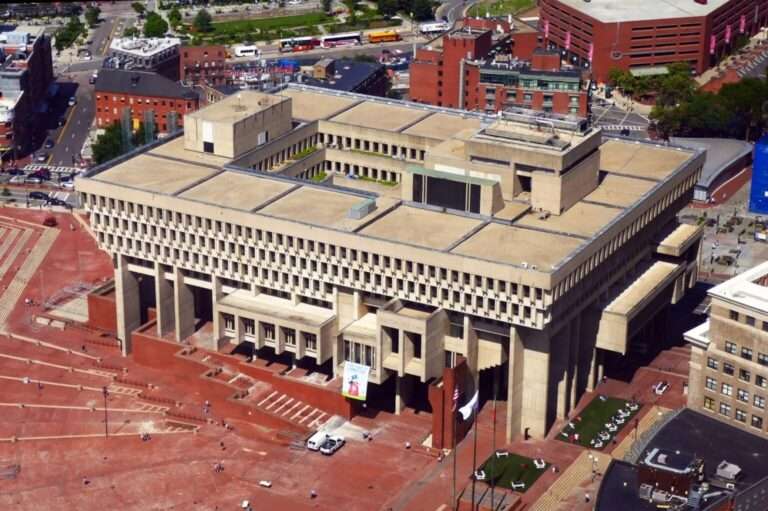 Aerial view of Boston City Hall showcasing its massive rectangular Brutalist structure, inner courtyard, tiered concrete cantilevers, and the vast red-brick City Hall Plaza surrounding it.
