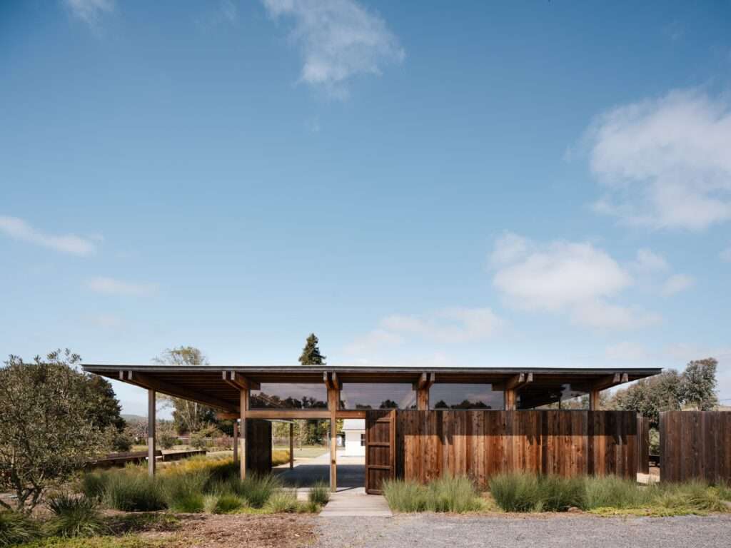 Exterior front view of the Live Oak Farm Pavilion featuring a floating roof design, wooden vertical cladding, clear clerestory windows, and a wooden footbridge pathway surrounded by native grasses.