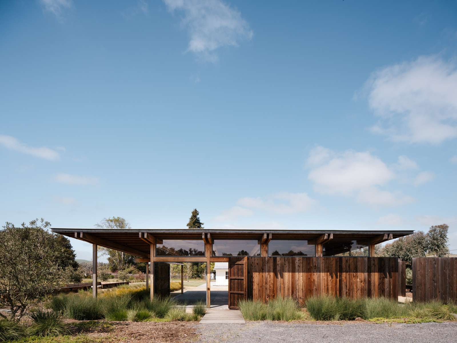 Exterior front view of the Live Oak Farm Pavilion featuring a floating roof design, wooden vertical cladding, clear clerestory windows, and a wooden footbridge pathway surrounded by native grasses.