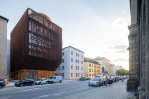 Exterior view of the five-story Kinderoase an der TUM daycare center in Munich, showcasing its textured timber facade and lower cantilevered structure integrated into an urban streetscape.