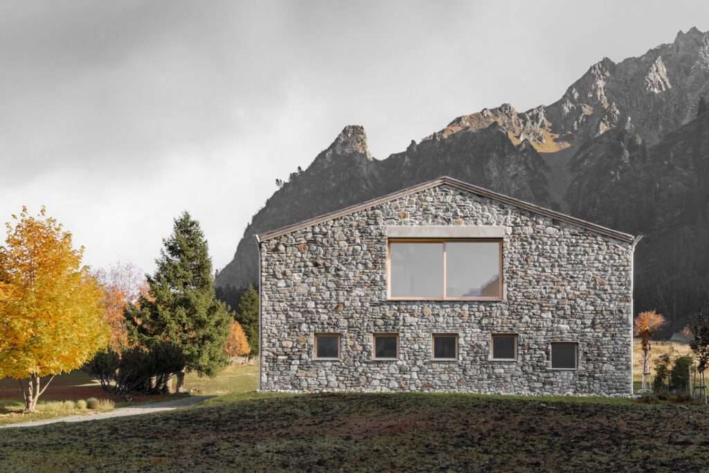 A two-story contemporary Alpine building with a full facade of locally sourced natural stone and a simple gabled roof, set against rugged mountains. Large windows on the upper floor frame the view.