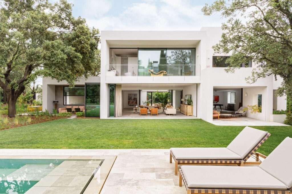 Rear facade of the renovated 1960s house with large sliding glass doors, a manicured lawn, an outdoor swimming pool, and lounge chairs under mature trees.