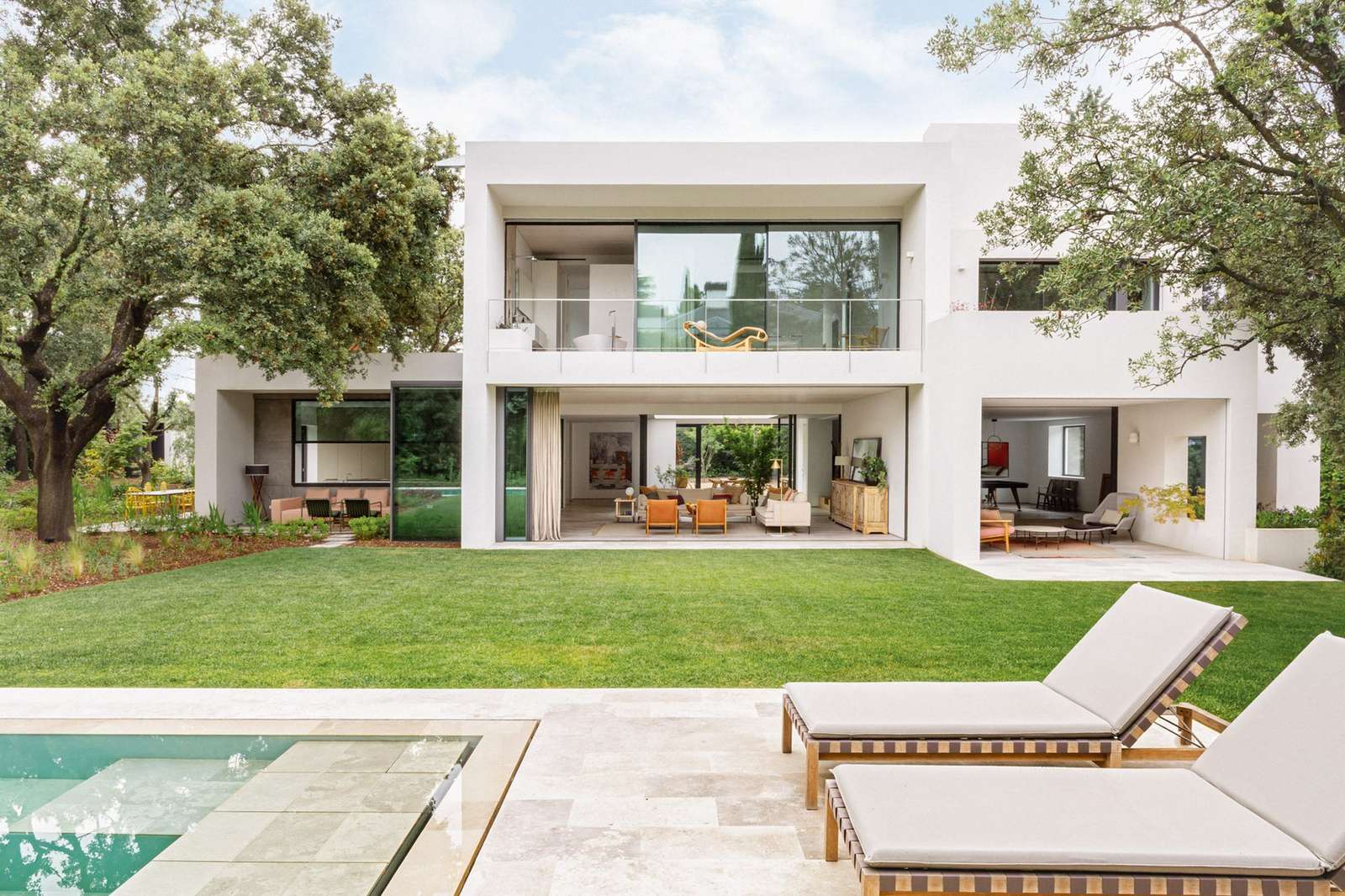Rear facade of the renovated 1960s house with large sliding glass doors, a manicured lawn, an outdoor swimming pool, and lounge chairs under mature trees.