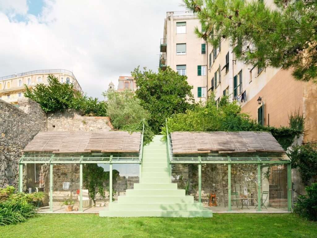 A front-facing photograph taken during the daytime shows a small, green-painted greenhouse with two sloping glass roofs flanking a central staircase made of the same green material. The structures are built against a rugged, historical stone wall, with lush trees and dense vegetation growing behind them. Behind the greenery, tall, older multi-story apartment buildings with green shutters are visible, one featuring clothes hanging to dry. The foreground is a well-maintained green lawn.