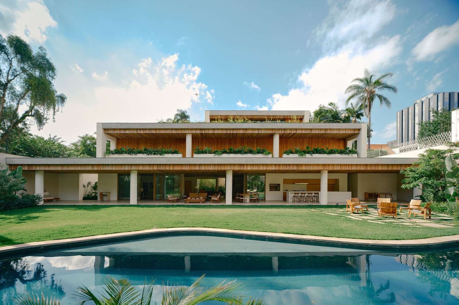 Rear facade of the two-story Jacarezinho II House with long verandas, a continuous wood slat ceiling, a lush green lawn, and a swimming pool reflecting the cloudy blue sky.