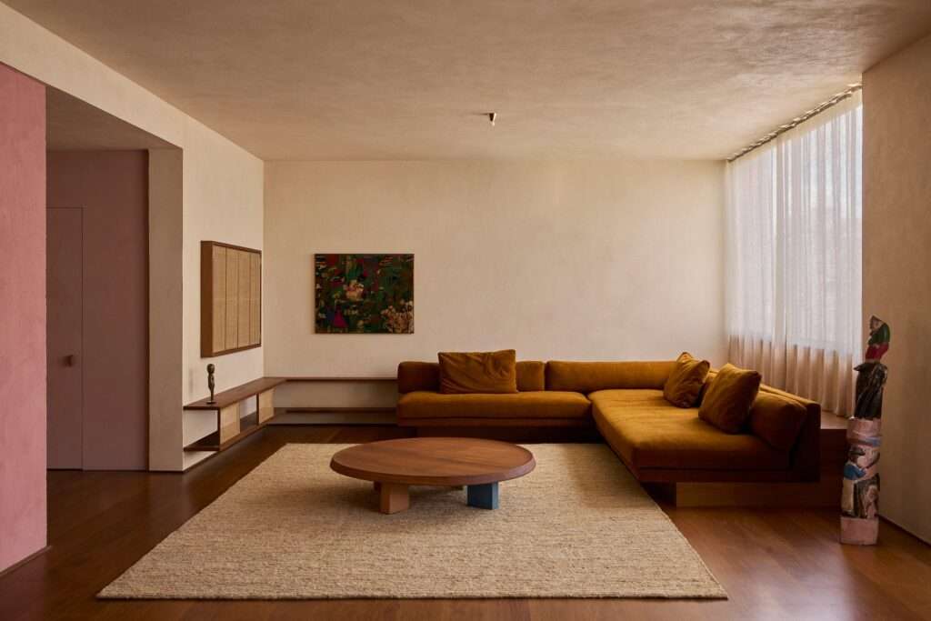 Panoramic view of an open-plan living area in a renovated Higienópolis apartment, featuring a large mustard-yellow velvet sectional sofa, a central round wooden coffee table on a jute rug, rich wood flooring, and large windows with sheer curtains. A pink-painted entryway wall is visible to the left.