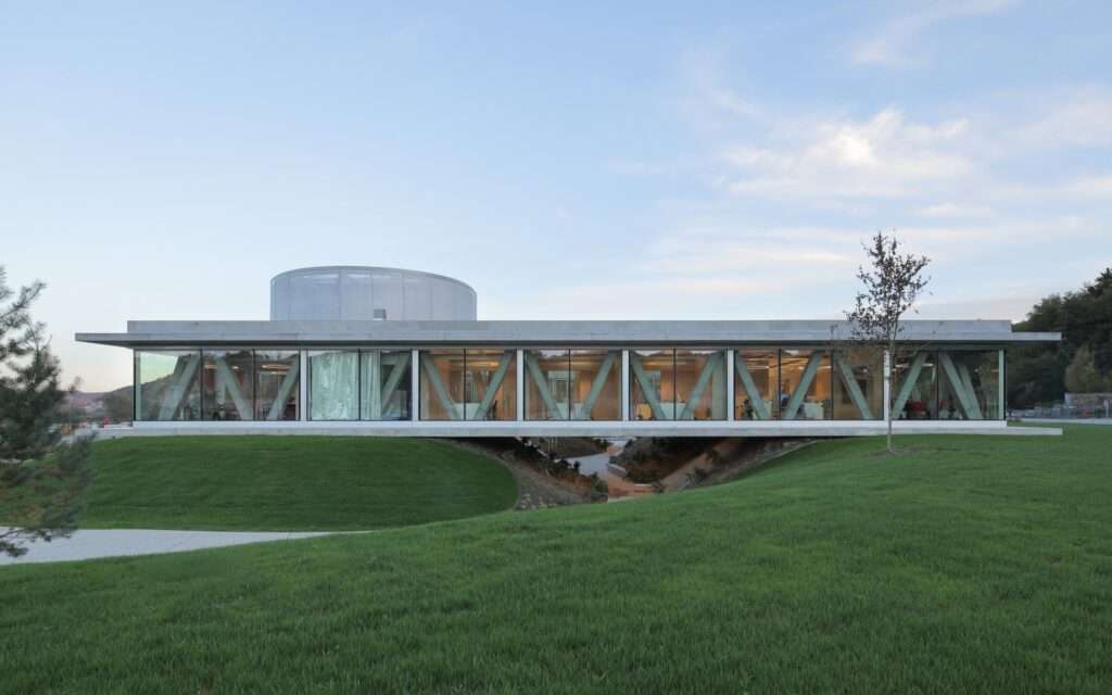 Exterior view of the elevated square volume of the Pina Astra office building resting atop grassy artificial hills with V-shaped concrete columns and full-height glass facades.