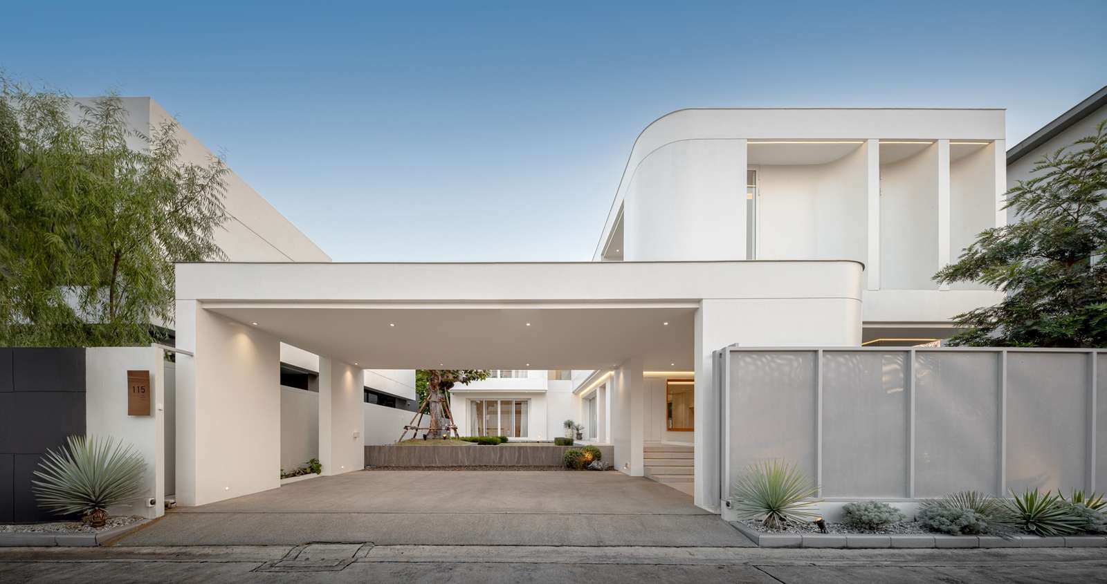 Front elevation view of JNG House showing a modern white minimalist two-story villa with a spacious integrated carport, curved architectural lines, and private boundary screen walls.