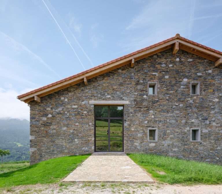 Front facade of Casa Basque House featuring restored original stone masonry walls and a contemporary large glass entryway under a gabled terracotta tile roof.
