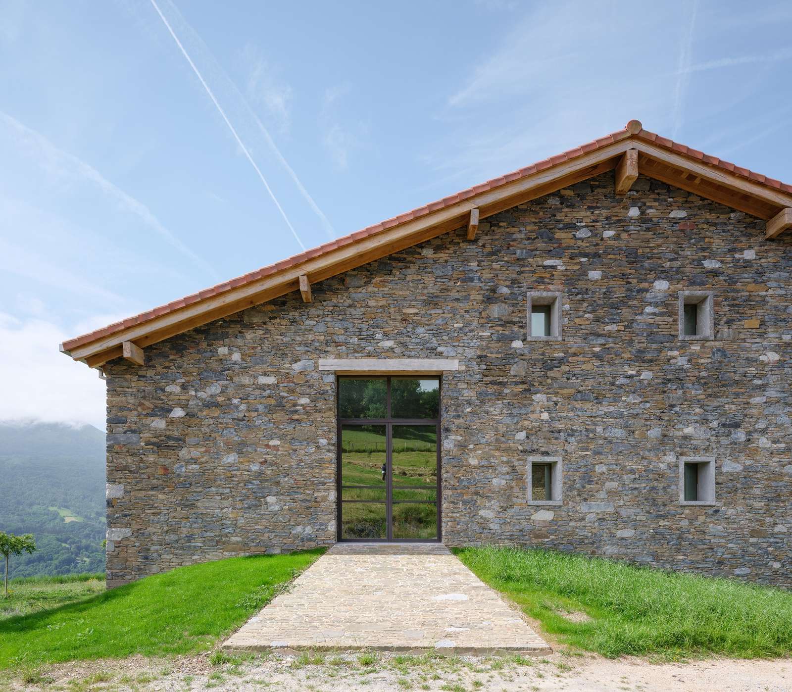 Front facade of Casa Basque House featuring restored original stone masonry walls and a contemporary large glass entryway under a gabled terracotta tile roof.