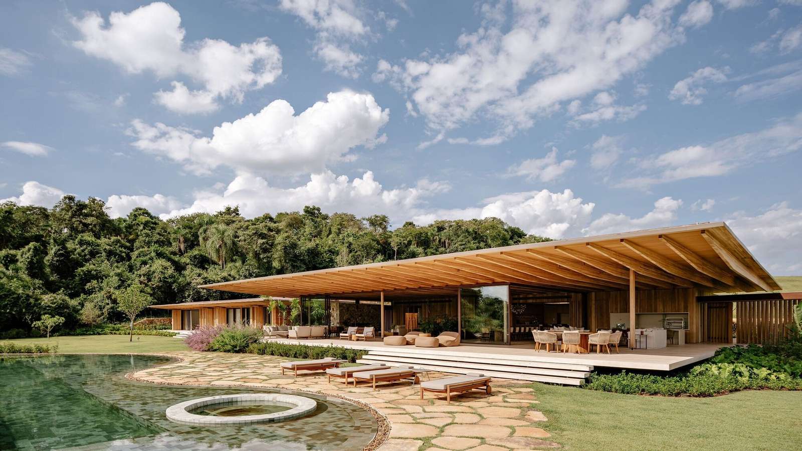 Exterior wide angle shot of AMS House showing a modern one-story pavilion with a large cantilevered timber roof, stone patio, outdoor pool, and dense forest background.