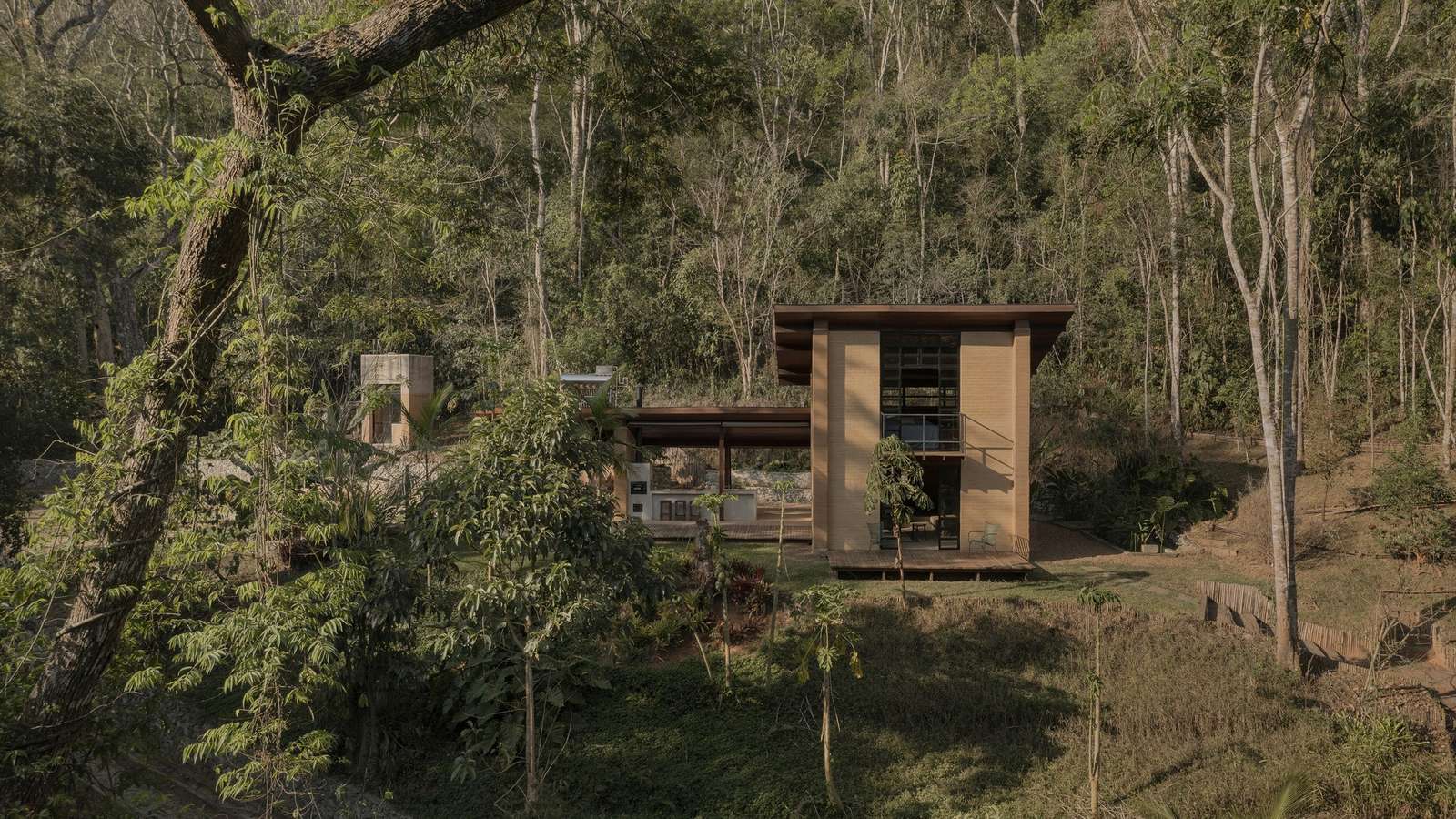 A rammed earth cabin nestled within a dense, lush green forest, viewed from a slightly elevated position across a grassy slope.
