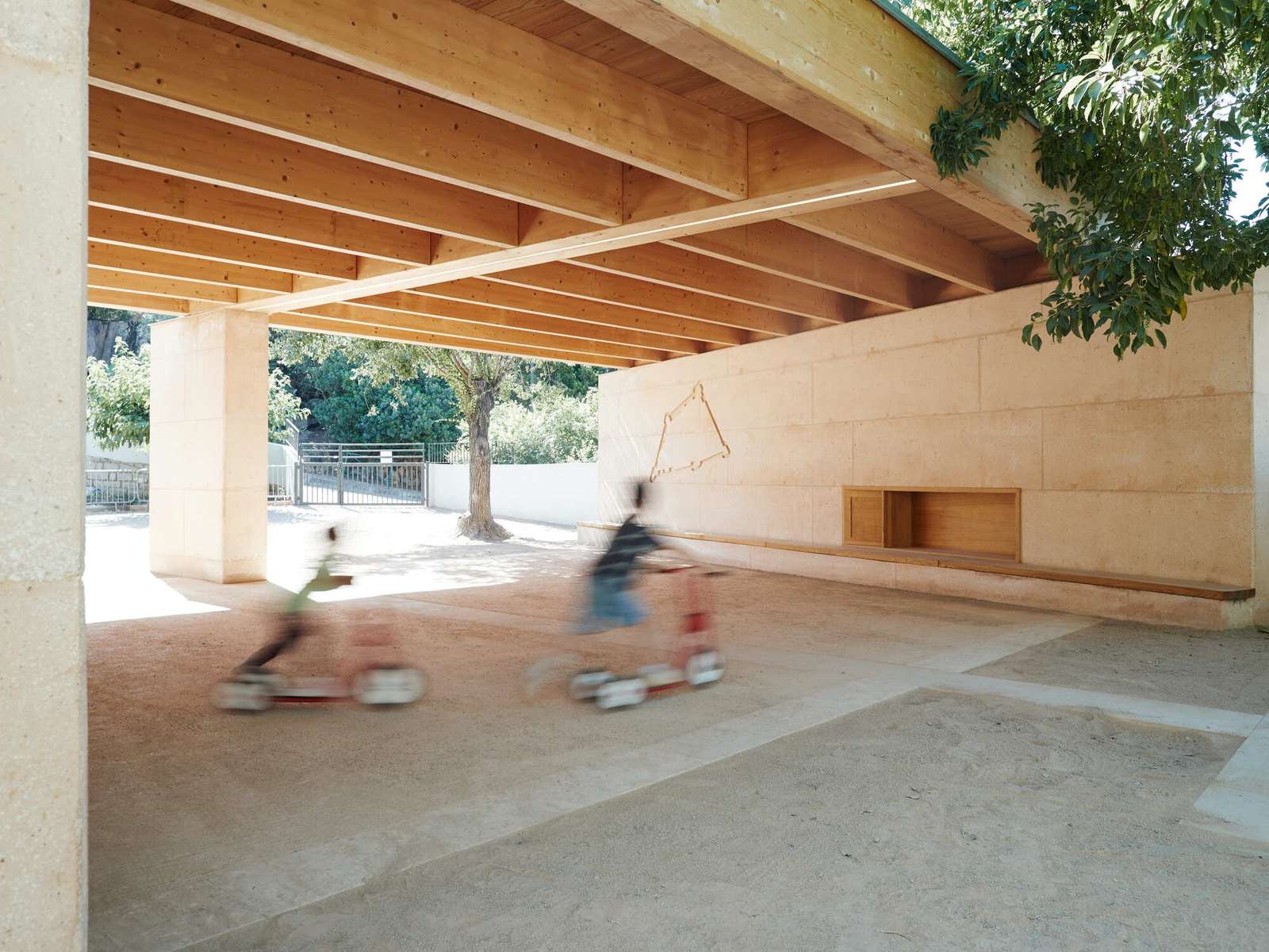 Two young children riding toy scooters beneath a large local wooden canopy at Josette Vincensini School courtyard next to a textured concrete wall.