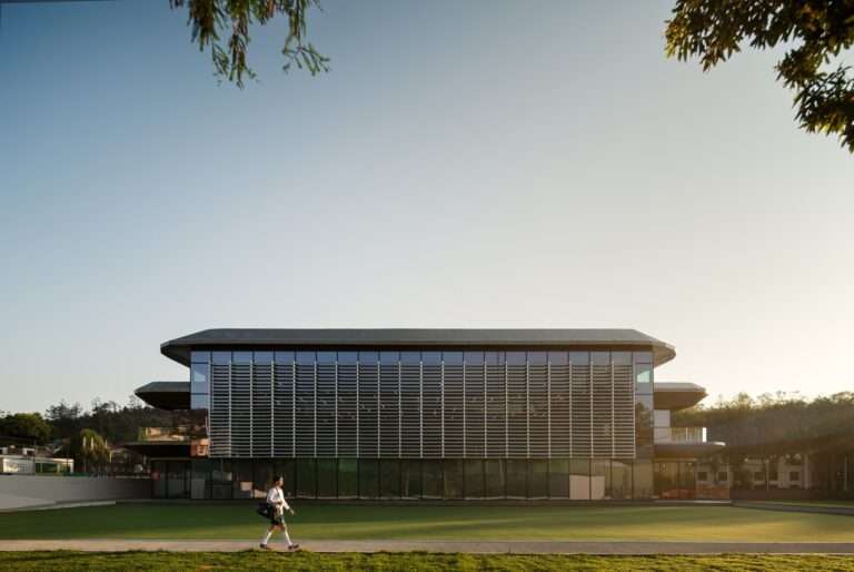 Modern three-story sports facility facade with extensive glass windows and a prominent horizontal louver system facing a green grass field under a clear blue sky.