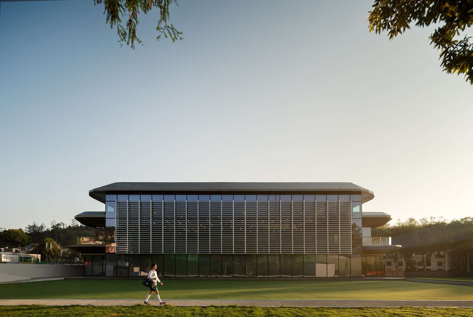 Modern three-story sports facility facade with extensive glass windows and a prominent horizontal louver system facing a green grass field under a clear blue sky.