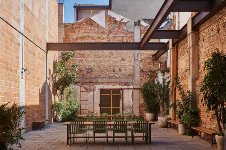 Adaptive reuse of Fábrica Bhering in Rio de Janeiro, Brazil. View of an outdoor courtyard with a communal dining table and green metal chairs, surrounded by exposed brick walls, steel beams, and potted plants. A red ladder leans against the wall. Photographed by Rafael Salim.