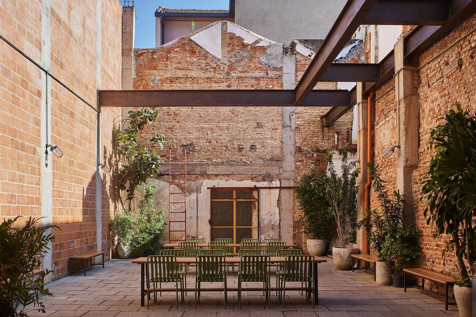 Adaptive reuse of Fábrica Bhering in Rio de Janeiro, Brazil. View of an outdoor courtyard with a communal dining table and green metal chairs, surrounded by exposed brick walls, steel beams, and potted plants. A red ladder leans against the wall. Photographed by Rafael Salim.