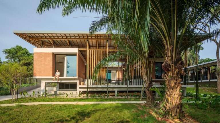 Exterior view of Anjung House showing compressed earth block walls, an elevated concrete platform, a prominent bamboo roof structure, and surrounding tropical palm trees.