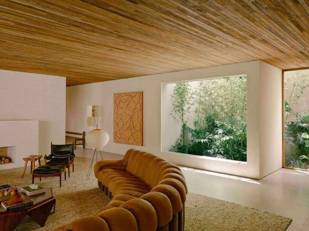 Minimalist living room interior of Jacarezinho II House featuring a curved modular camel-colored sofa, a warm wooden slat ceiling, and a large square window framing a lush pocket garden.