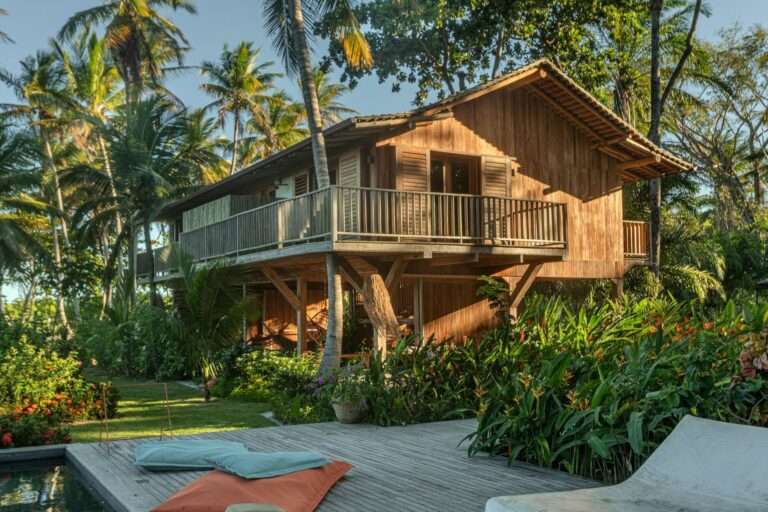 Two-story tropical wooden house with elevated balcony surrounded by palm trees and lush vegetation near a pool deck.