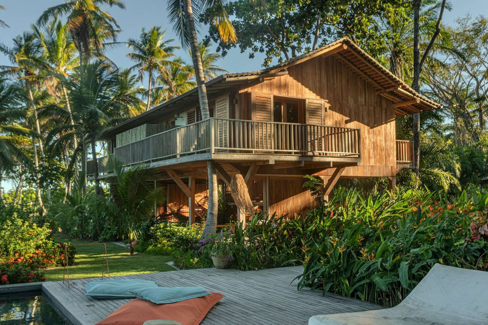 Two-story tropical wooden house with elevated balcony surrounded by palm trees and lush vegetation near a pool deck.