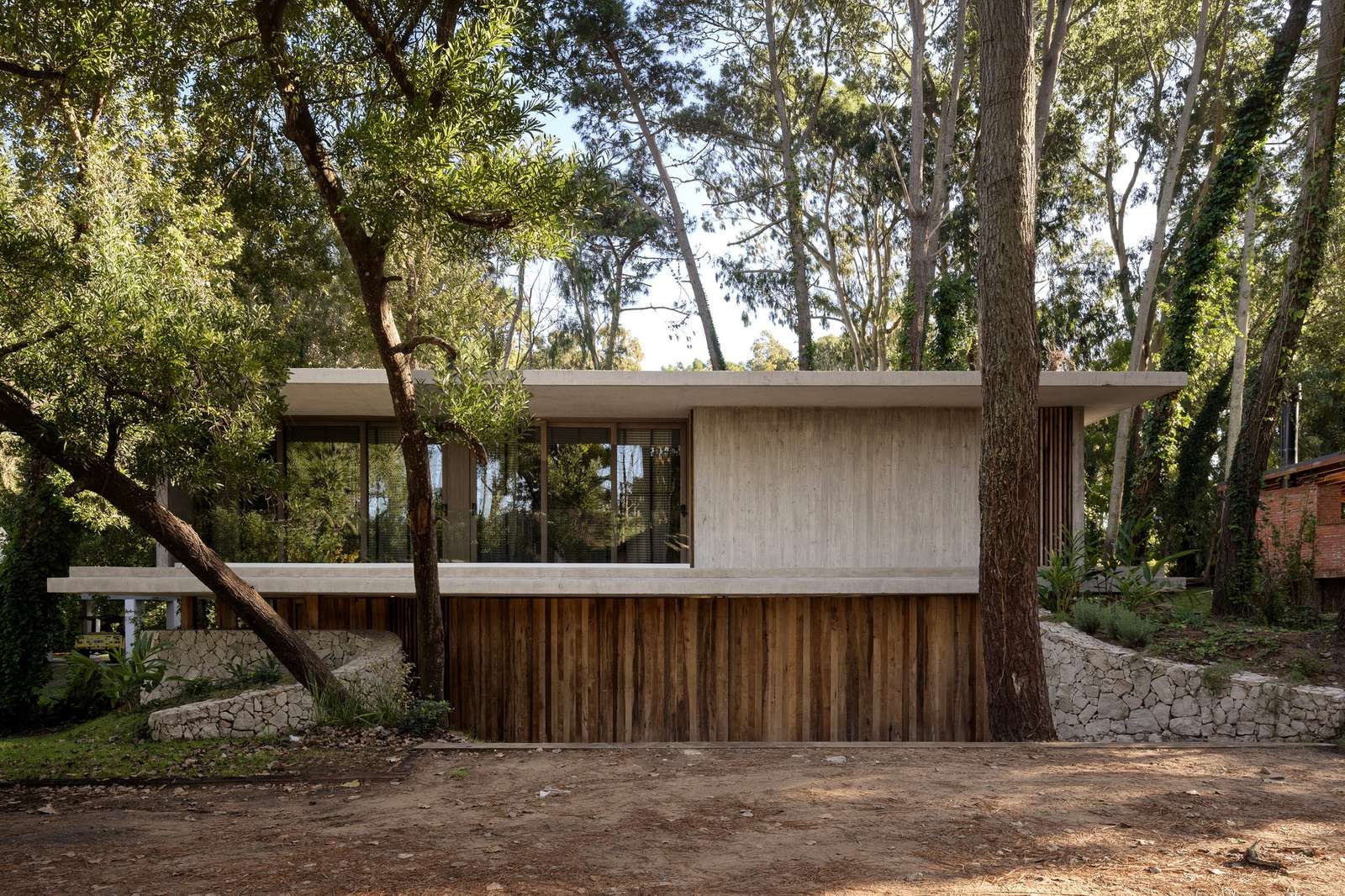 Exterior elevation of Maison A showing a horizontal concrete slab, vertical timber cladding, and a rustic stone foundation integrated with tall forest trees.