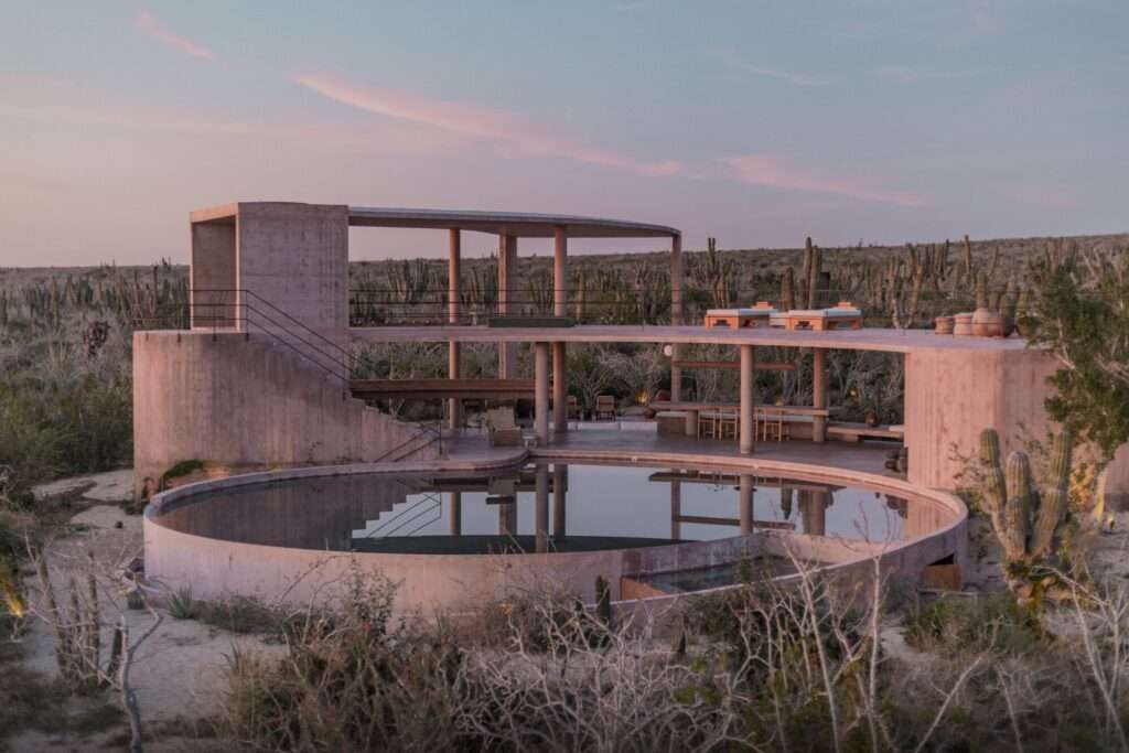 Exterior view of a modern desert house made of pigment-dyed pink concrete, featuring a multi-level structure with a large circular swimming pool at the base and a long ramp leading to a second-floor terrace.