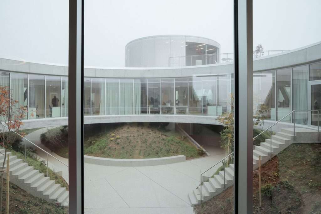 Looking through a glass window into a circular central courtyard surrounded by curved floor-to-ceiling glass walls, outdoor staircases, and planted slopes under a foggy sky.