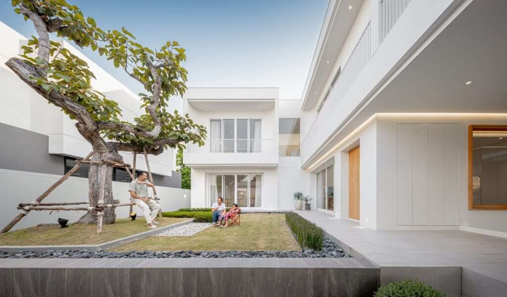 A family relaxing in the central green courtyard lawn of the JNG House, surrounded by white L-shaped building wings with large glass doors.