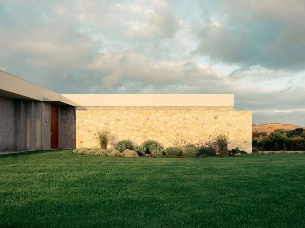 Textured stone retaining wall bordering green lawn with the low-profile concrete architecture of Wildcoast House under a cloudy sky.