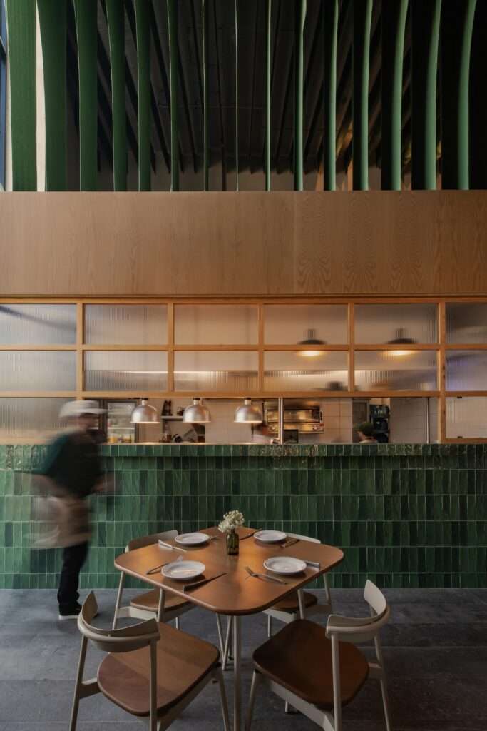 Open kitchen service counter clad in dark green glazed tiles with fluted glass windows, wooden cladding, and a set dining table in the foreground.