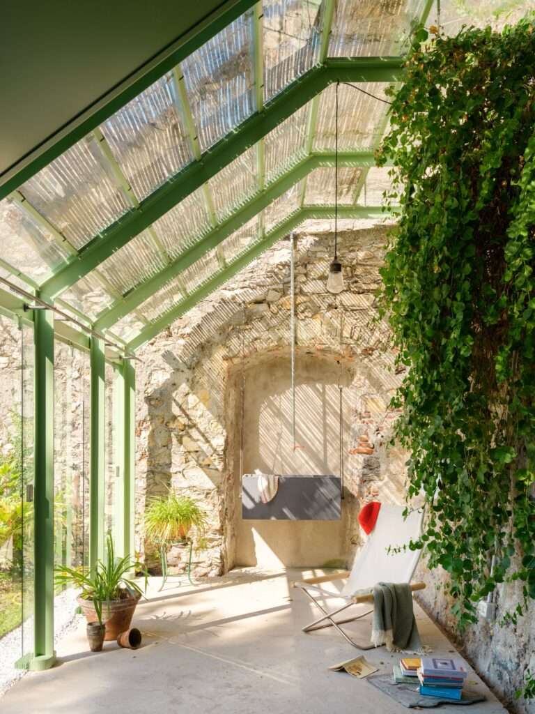 An interior photograph looking down the length of the greenhouse during daytime, showing a light-filled space with transparent glass walls and roof panels framed in light green metal. Sunlight streams in, casting patterned shadows. The space features a historical textured stone archway with a hanging planter and tools. Potted plants line the perimeter, including a prominent snake plant. Further down, there is a small seating area with a chair and books.