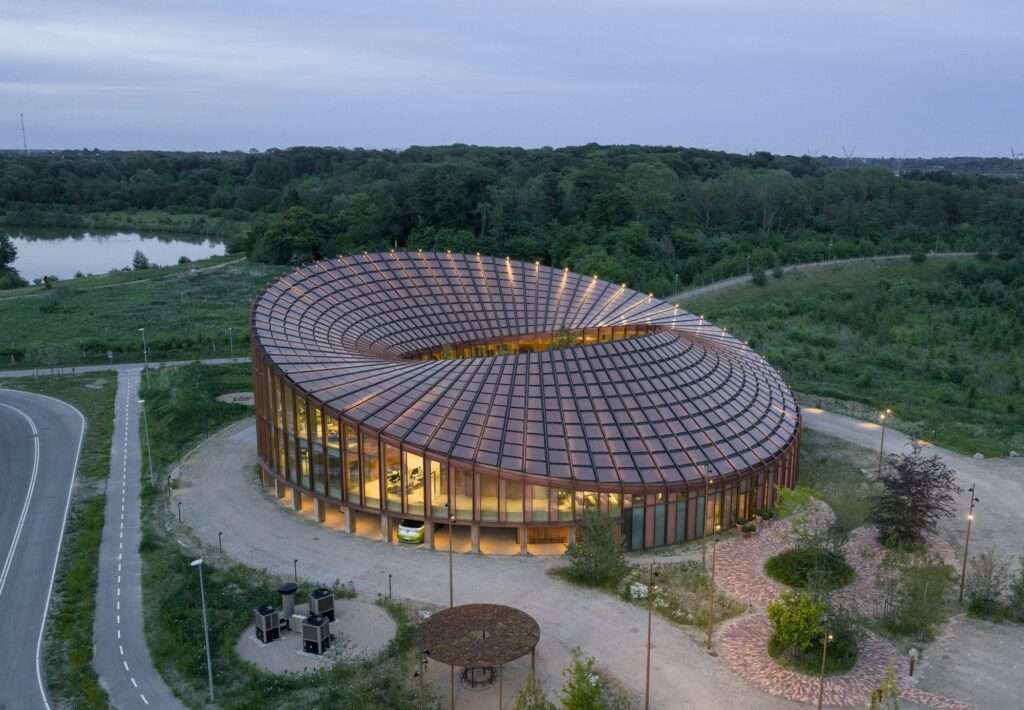 Aerial evening view of the circular Dymak HQ building featuring a sweeping solar-paneled roof next to a lake and forest.