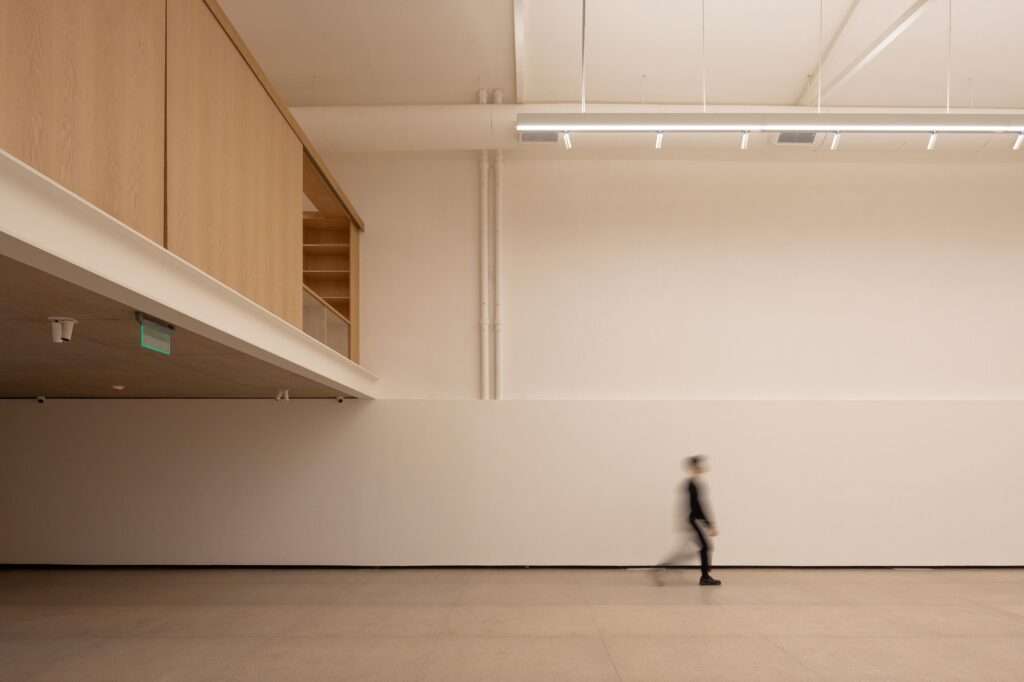 Minimalist double-height exhibition hall of the Larivière Foundation in La Boca, showing a warm oak wood mezzanine on the left, white walls, industrial lighting, and a person walking in motion blur.